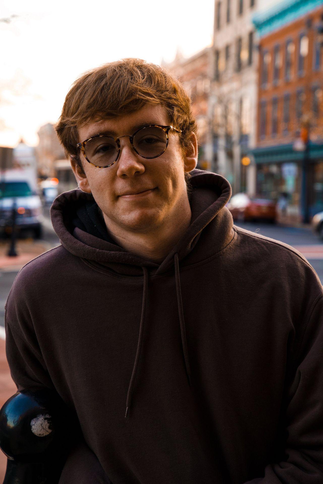 Person wearing round glasses and a hoodie standing on a street with buildings and vehicles in the background.