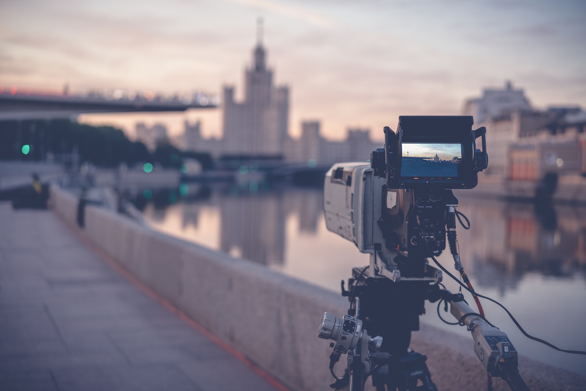 A professional video camera stands on a tripod recording the city and the river at sunrise, documentary filming in Moscow